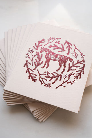 Photo of a holiday greeting card featuring a foil stamped design of a horse amongst pine boughs and holly branches.