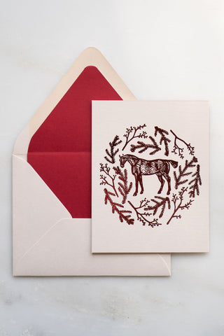 Photo of a holiday greeting card featuring a foil stamped design of a horse amongst pine boughs and holly branches.