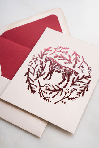 Photo of a holiday greeting card featuring a foil stamped design of a horse amongst pine boughs and holly branches.