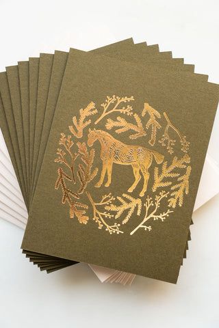 Photo of a holiday greeting card featuring a foil stamped design of a horse amongst pine boughs and holly branches.