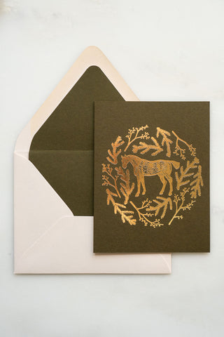 Photo of a holiday greeting card featuring a foil stamped design of a horse amongst pine boughs and holly branches.