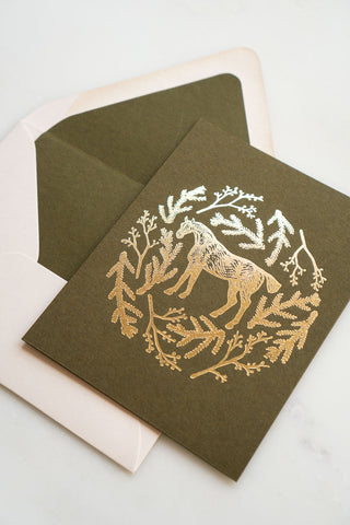 Photo of a holiday greeting card featuring a foil stamped design of a horse amongst pine boughs and holly branches.