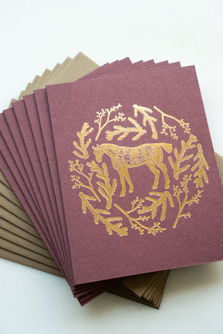 Photo of a holiday greeting card featuring a foil stamped design of a horse amongst pine boughs and holly branches.