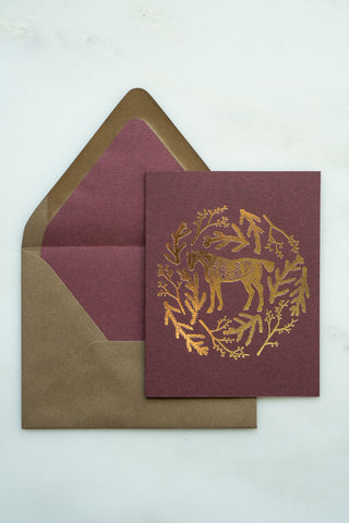 Photo of a holiday greeting card featuring a foil stamped design of a horse amongst pine boughs and holly branches.