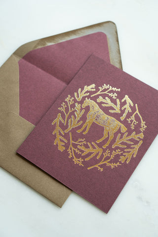 Photo of a holiday greeting card featuring a foil stamped design of a horse amongst pine boughs and holly branches.