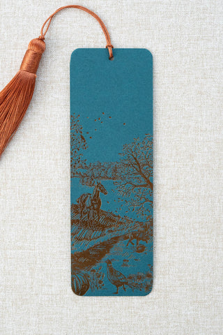 A foil stamped bookmark featuring a countryside autumn scene with a horse, fox, pumpkins, pheasant, and crow. Printed in bronze foil on a dark teal card stock and finished with a deep orange tassel.