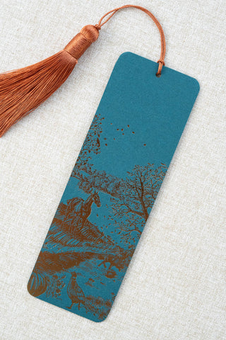 A foil stamped bookmark featuring a countryside autumn scene with a horse, fox, pumpkins, pheasant, and crow. Printed in bronze foil on a dark teal card stock and finished with a deep orange tassel.