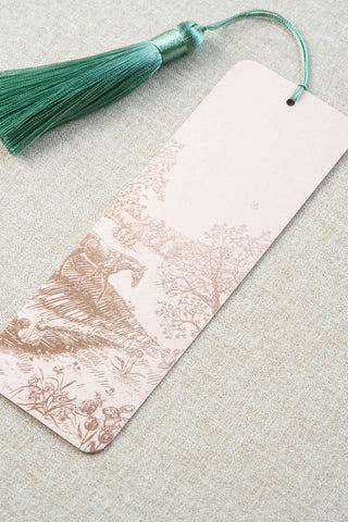 A foil stamped bookmark featuring a countryside spring scene with a horse, swan, rabbits, butterflies and flowers. Printed in rose gold foil on a light peach pink card stock and finished with a light turquoise tassel.