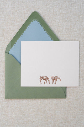 A cream correspondence card featuring two horse illustrations foil stamped in rose gold. Accompanying medium green envelope is lined with a scalloped-edge dusty blue envelope liner.