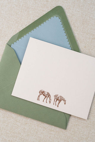 A cream correspondence card featuring two horse illustrations foil stamped in rose gold. Accompanying medium green envelope is lined with a scalloped-edge dusty blue envelope liner.