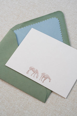 A cream correspondence card featuring two horse illustrations foil stamped in rose gold. Accompanying medium green envelope is lined with a scalloped-edge dusty blue envelope liner.