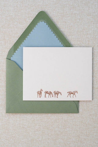 A cream correspondence card featuring four horse illustrations foil stamped in rose gold. Accompanying medium green envelope is lined in a scalloped-edge dusty blue envelope liner.