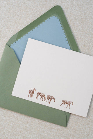 A cream correspondence card featuring four horse illustrations foil stamped in rose gold. Accompanying medium green envelope is lined in a scalloped-edge dusty blue envelope liner.