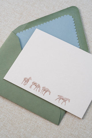 A cream correspondence card featuring four horse illustrations foil stamped in rose gold. Accompanying medium green envelope is lined with a scalloped-edge dusty blue envelope liner.