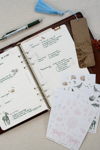 Washi paper stickers used in a Filofax planner page layout. Planner laid open to a two page weekly planner spread. Washi paper sticker sheets featuring delicate illustrations of horses, wildlife and flowers lay over a bottom corner of the planner spread, a foil stamped horse motif bookmark marks the page and a pen sits above.