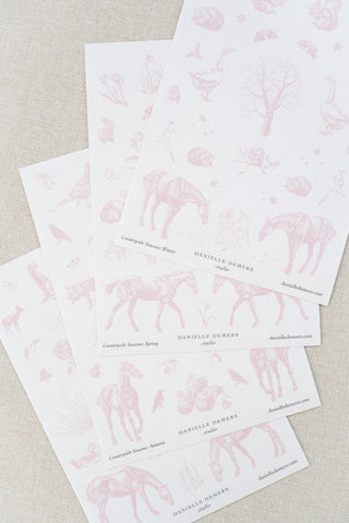 Four sheets of Danielle Demers washi paper sticker sheets fanned out on a light beige textured fabric background. The sticker sheets feature delicate pink illustrations of horses, countryside animals, and flowers.