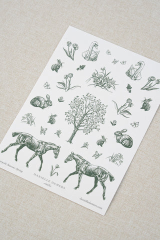 Danielle Demers washi paper sticker sheet on a light beige textured fabric background. The sticker sheet features delicate forest green illustrations of horses, countryside animals, and flowers.