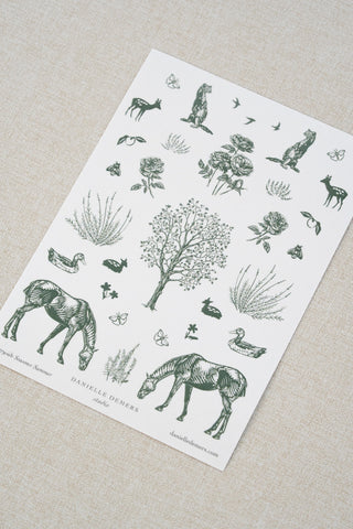 Danielle Demers washi paper sticker sheet on a light beige textured fabric background. The sticker sheet features delicate forest green illustrations of horses, countryside animals, and flowers.