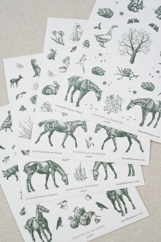Four sheets of Danielle Demers washi paper sticker sheets fanned out on a light beige textured fabric background. The sticker sheets feature delicate forest green illustrations of horses, countryside animals, and flowers.