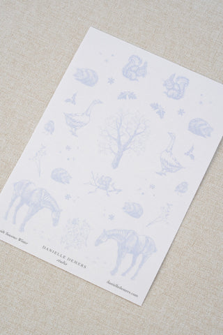 Danielle Demers washi paper sticker sheet on a light beige textured fabric background. The sticker sheet features delicate periwinkle blue illustrations of horses, countryside animals, and flowers.