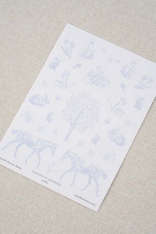 Danielle Demers washi paper sticker sheet on a light beige textured fabric background. The sticker sheet features delicate periwinkle blue illustrations of horses, countryside animals, and flowers.