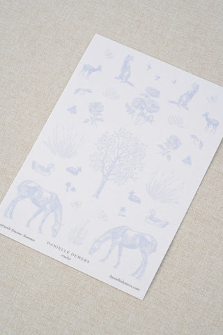 Danielle Demers washi paper sticker sheet on a light beige textured fabric background. The sticker sheet features delicate periwinkle blue illustrations of horses, countryside animals, and flowers.