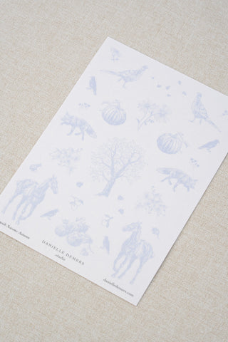 Danielle Demers washi paper sticker sheet on a light beige textured fabric background. The sticker sheet features delicate periwinkle blue illustrations of horses, countryside animals, and flowers.