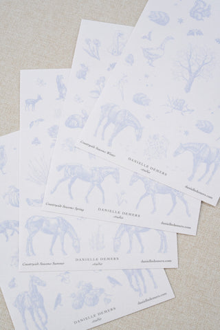 Four sheets of Danielle Demers washi paper sticker sheets fanned out on a light beige textured fabric background. The sticker sheets feature delicate periwinkle blue illustrations of horses, countryside animals, and flowers.