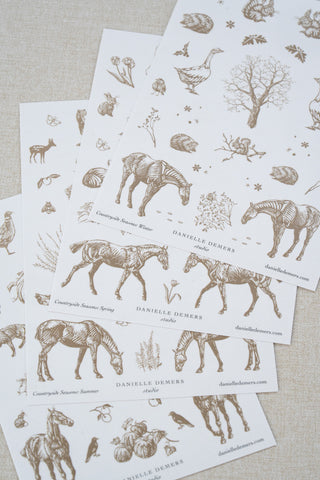Four sheets of Danielle Demers washi paper sticker sheets fanned out on a light beige textured fabric background. The sticker sheets feature delicate taupe brown illustrations of horses, countryside animals, and flowers.