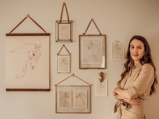 Photo of equine artist and surface pattern designer Danielle Demers posing with a gallery wall of framed prints of her horse drawings. Photo by Nicole Gearty.