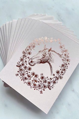 Photo of a set of 8 rich copper metallic foil stamped note cards, featuring a drawing of a horse in a wreath of cosmos flowers, bees and butterflies, on cream card stock by equine artist Danielle Demers.