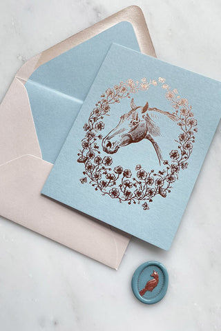Photo of a rich copper metallic foil stamped note card, featuring a drawing of a horse in a wreath of cosmos flowers, bees and butterflies, on pale blue 111lb card stock by equine artist Danielle Demers.