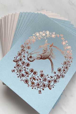 Photo of a set of 8 rich copper metallic foil stamped note cards, featuring a drawing of a horse in a wreath of cosmos flowers, bees and butterflies, on pale blue 111lb card stock by equine artist Danielle Demers.