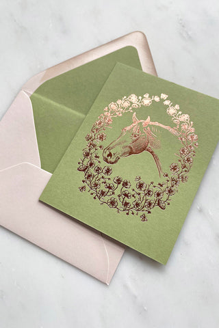 Photo of a rich copper metallic foil stamped note card, featuring a drawing of a horse in a wreath of cosmos flowers, bees and butterflies, on spring green 111lb card stock by equine artist Danielle Demers.
