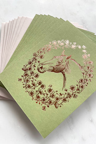 Photo of a set of 8 rich copper metallic foil stamped note cards, featuring a drawing of a horse in a wreath of cosmos flowers, bees and butterflies, on spring green 111lb card stock by equine artist Danielle Demers.